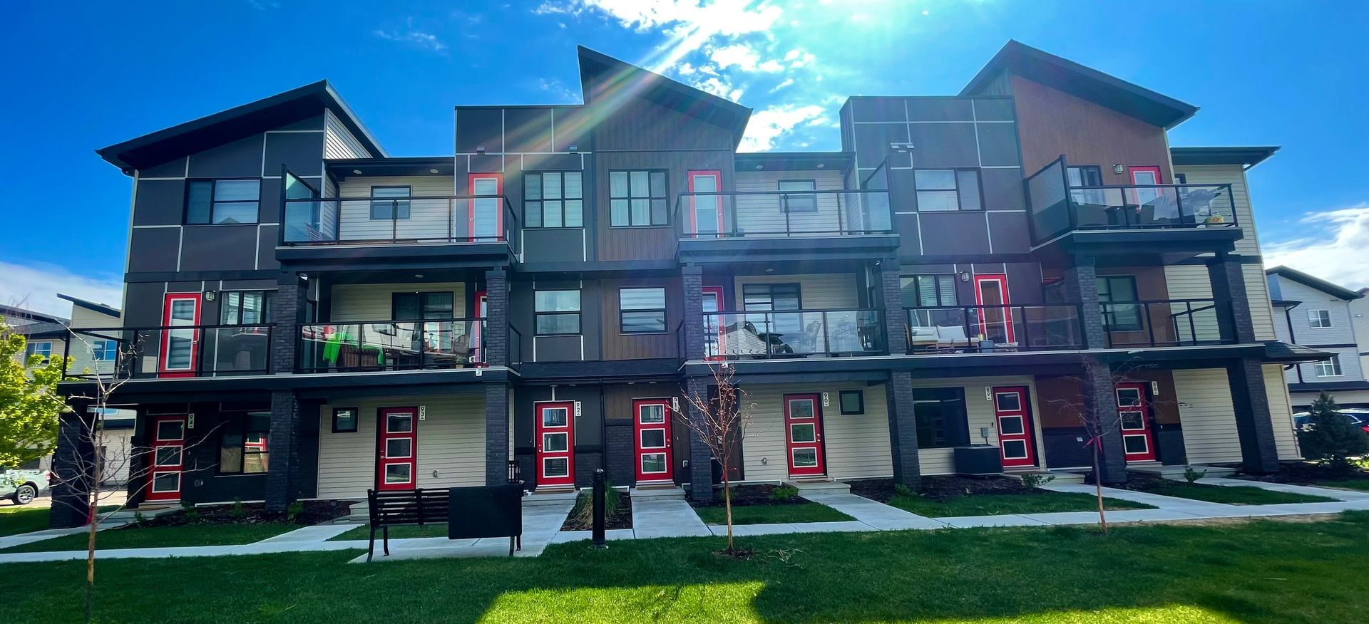 Modern three-story townhouses with red doors, balconies, and dark siding against a blue sky.