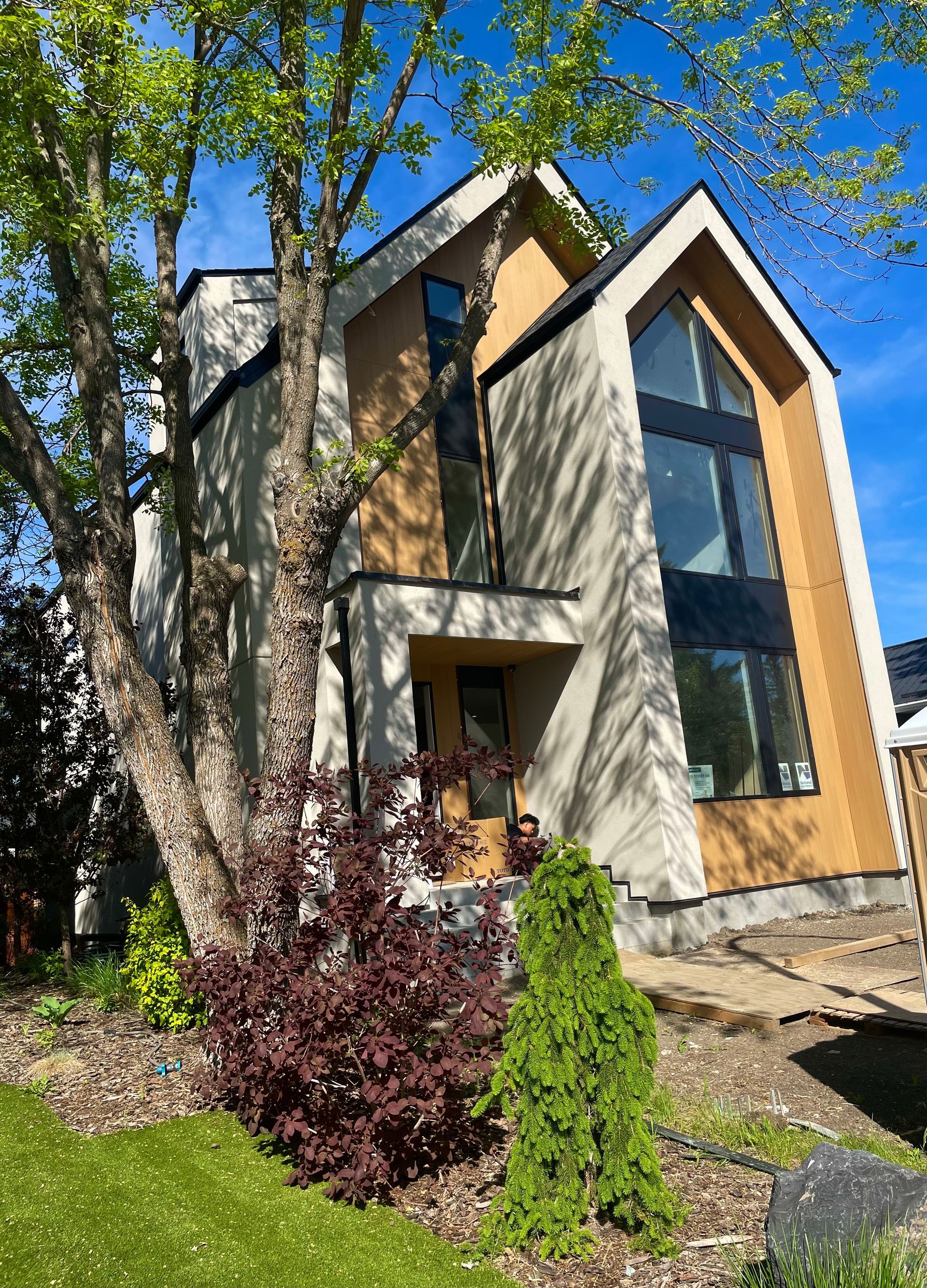Modern two-story house with large windows, beige and brown siding, and a front yard with trees.