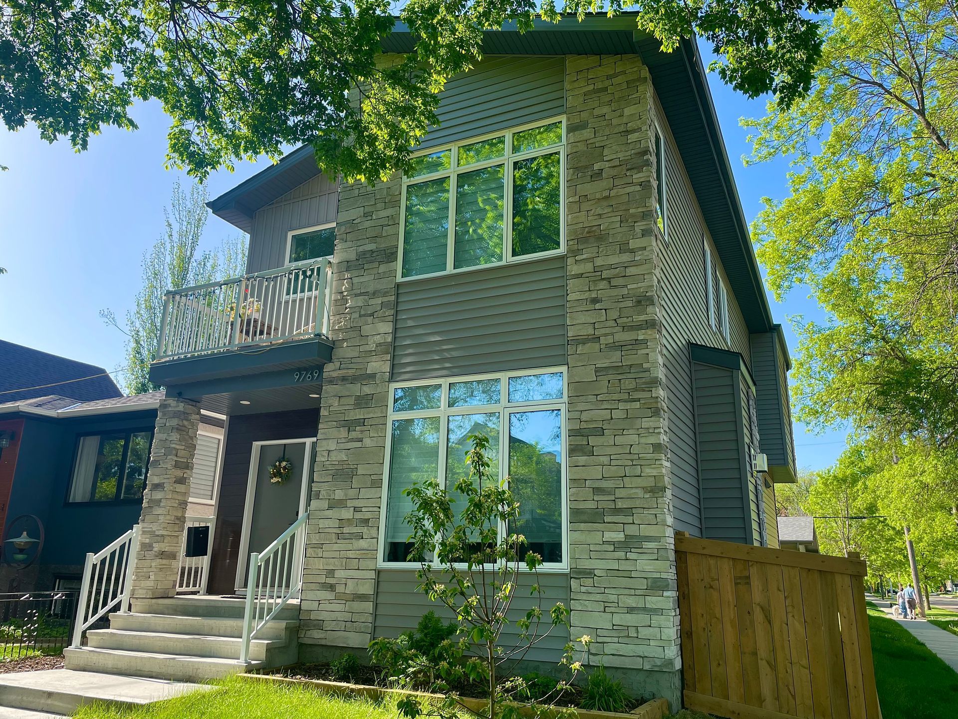 Two-story house with stone facade and large windows, fronted by a lawn and trees.
