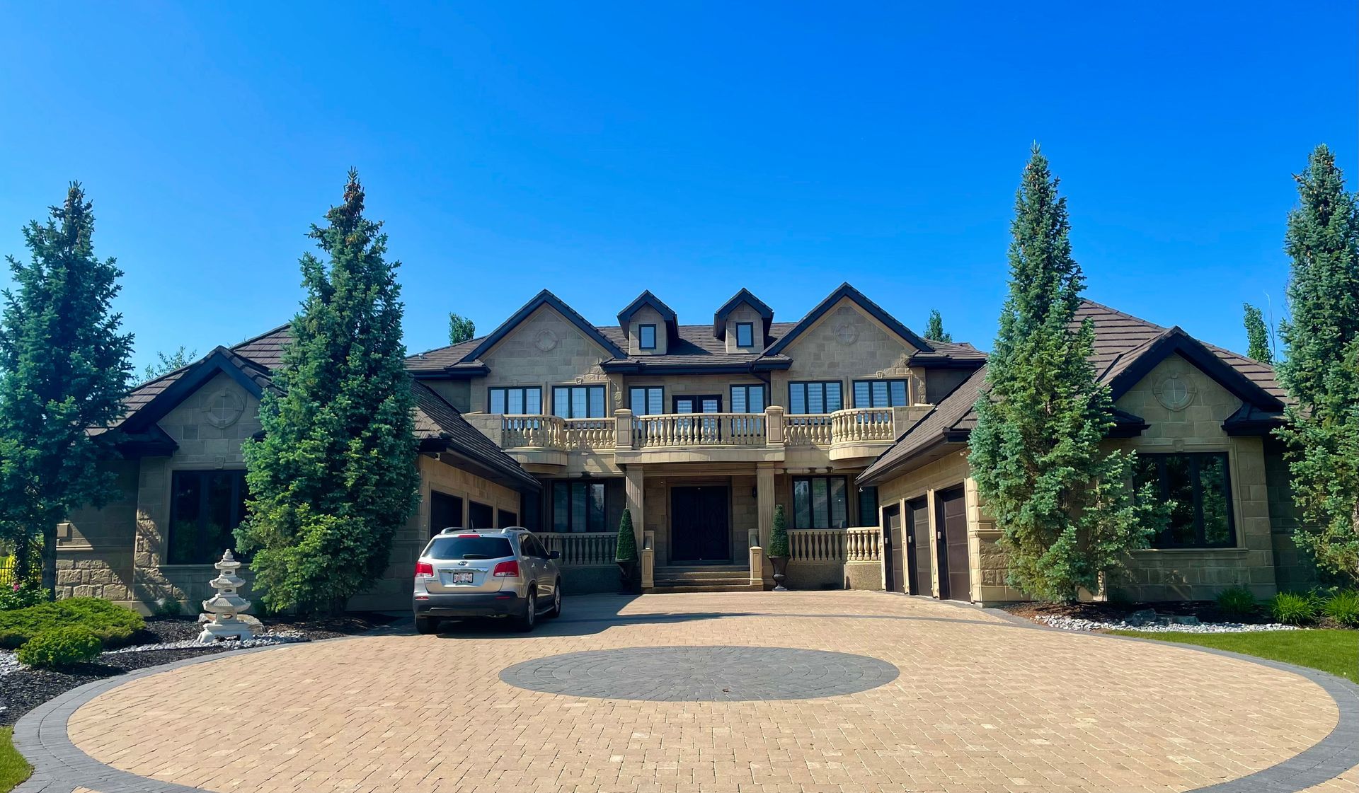 Large stone house with a circular driveway, car, and trees under a clear blue sky.