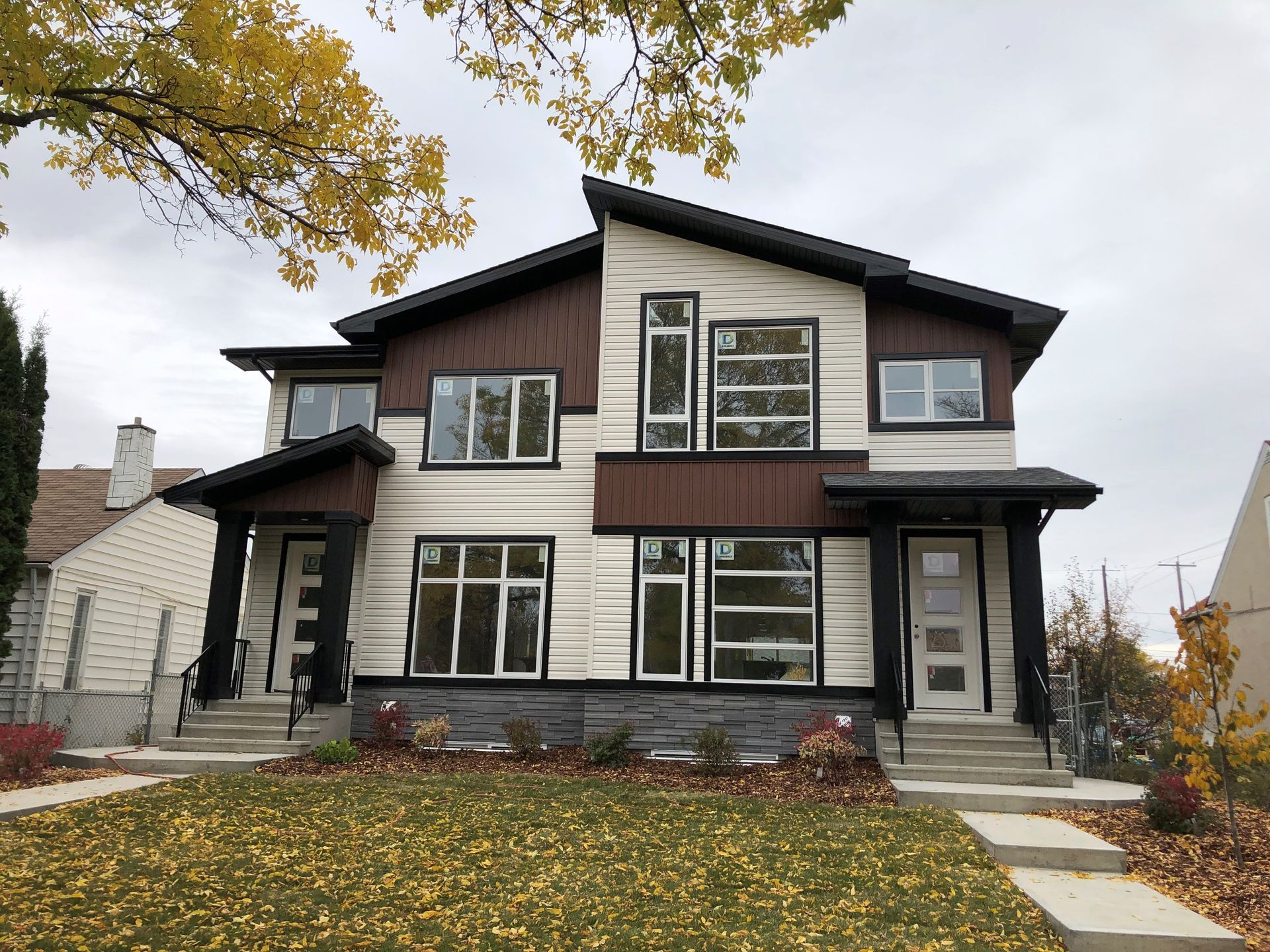 Two-story modern duplex with brown and white siding, black accents, and a grassy yard with fall leaves.