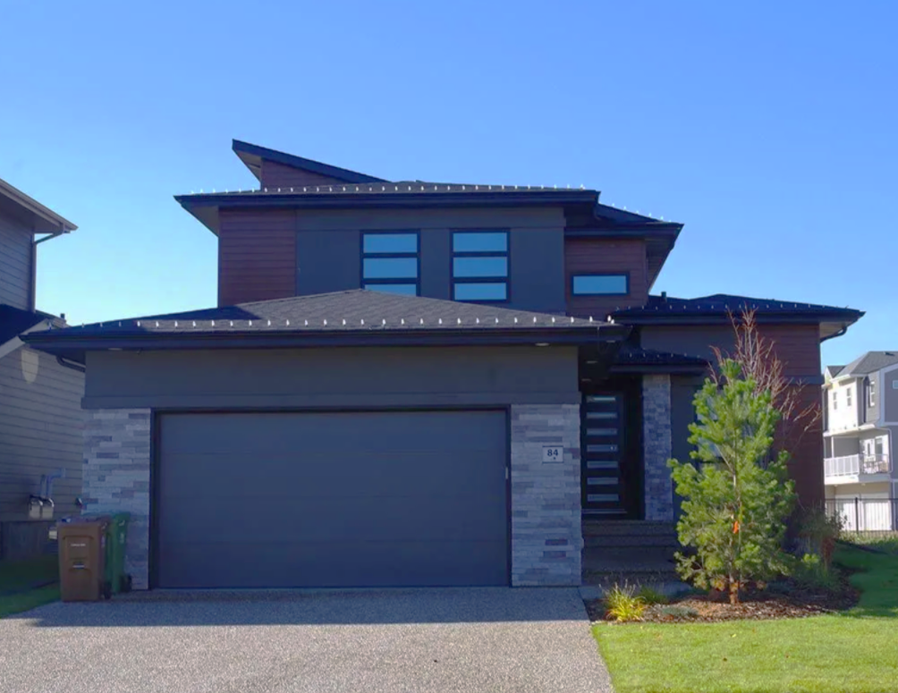 Modern two-story house with gray garage door, stone and wood siding, and a green lawn.