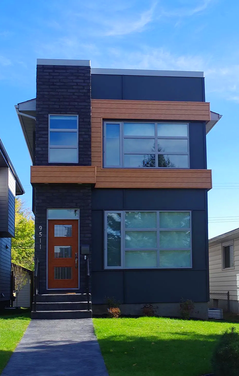 Modern two-story house with dark blue siding, wooden accents, and a bright orange door on a sunny day.
