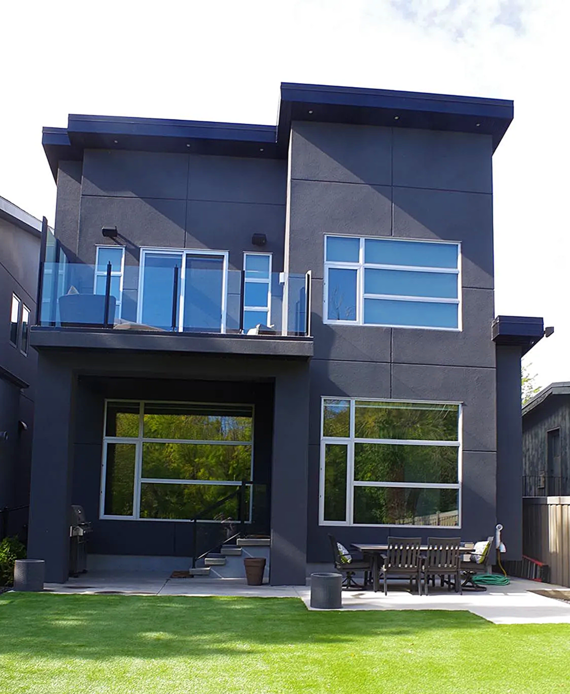 Modern two-story house with dark gray exterior, large windows, and glass balcony, set on a green lawn.