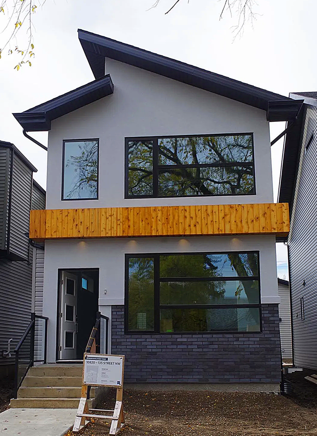 Modern two-story house with gray stucco and wood accents. Black windows and a sloped roof.