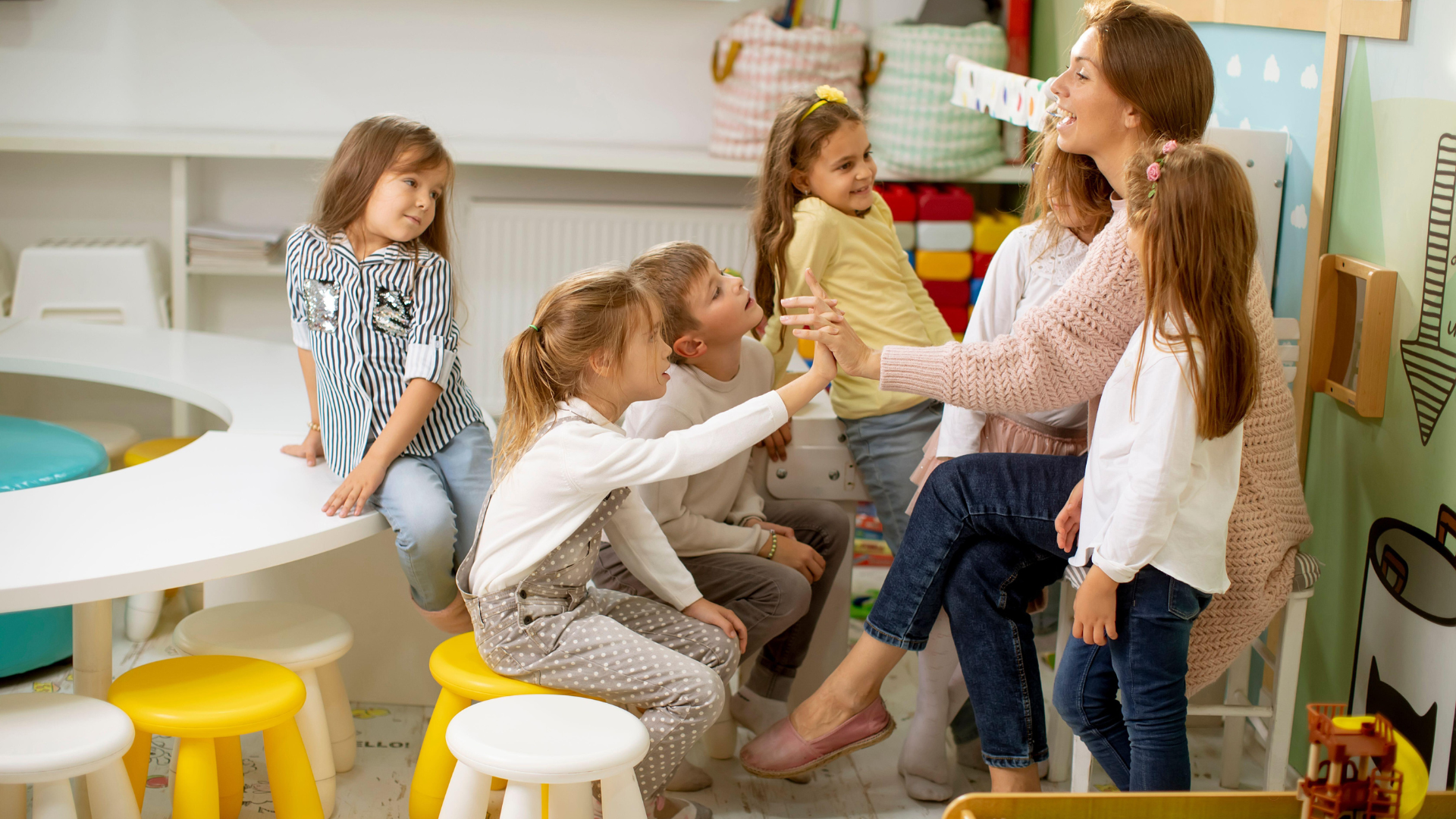 Group of young children interacting with a teacher in a daycare setting. The children are sitting at a table and engaged in a learning activity.