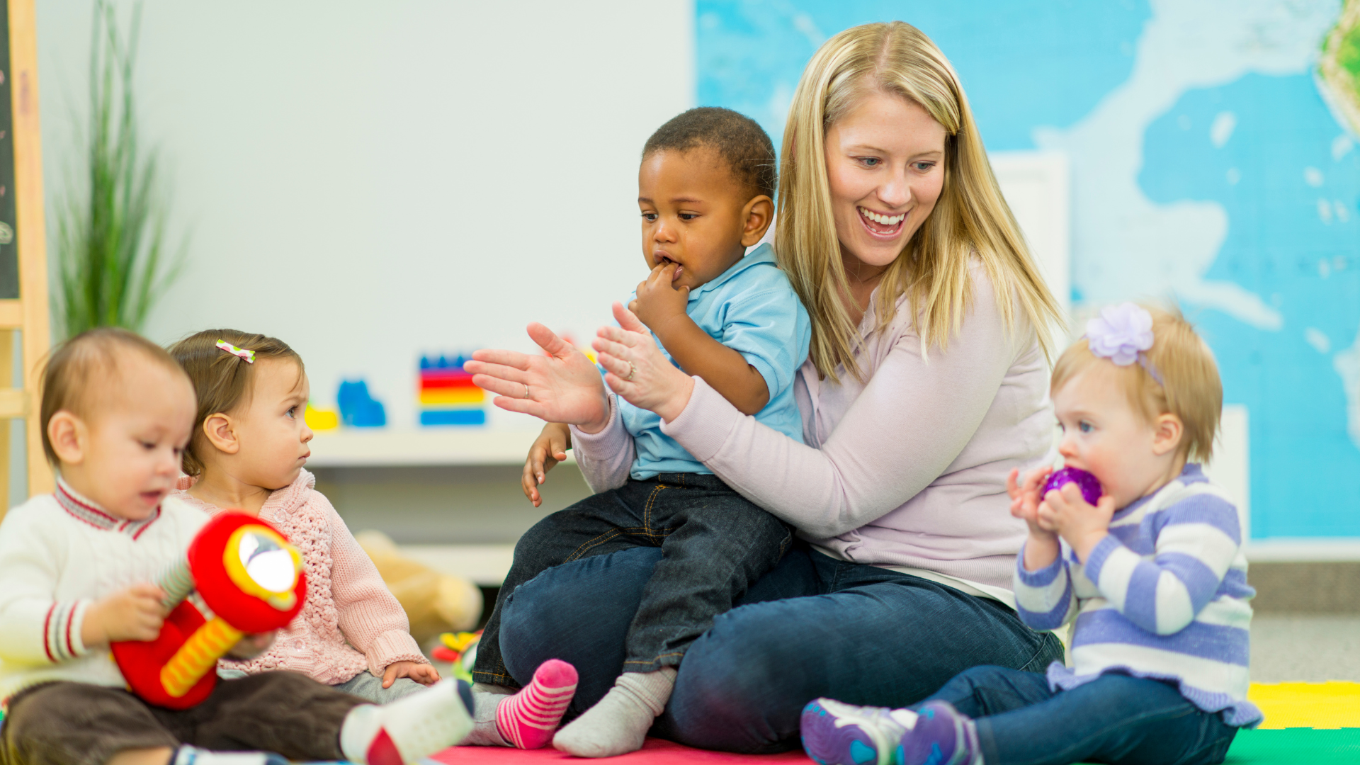 Infant and toddler playing with toys in a daycare classroom while being guided by a caregiver. The children are engaged in early learning activities.