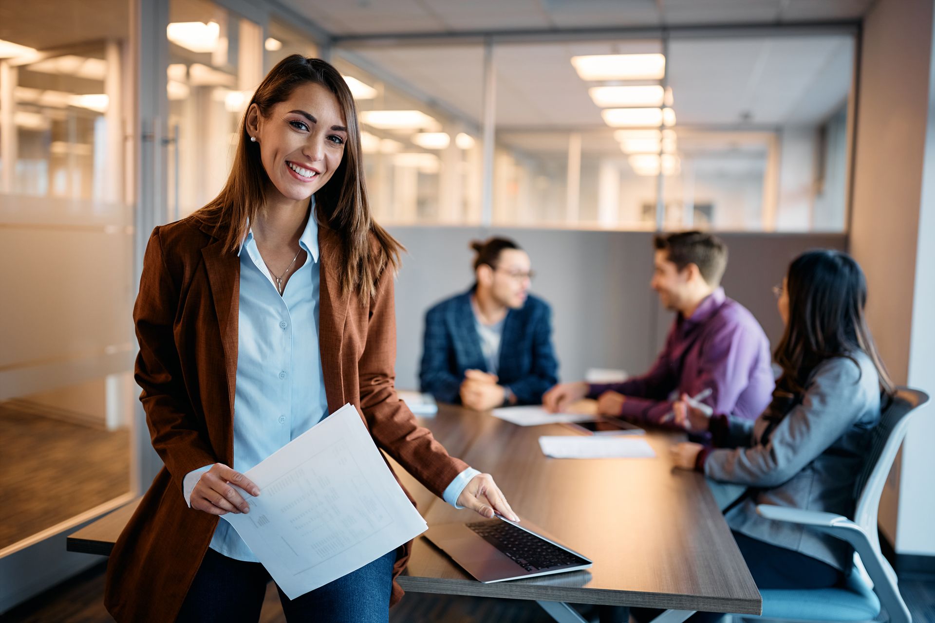 A woman is holding a piece of paper in front of a group of people sitting at a table.
