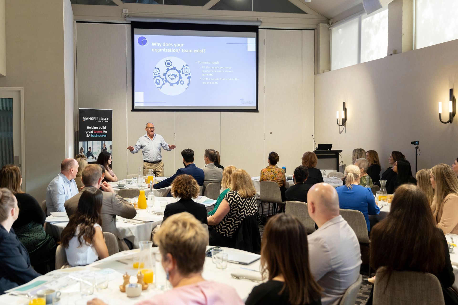 A group of people are sitting at tables in front of a projector screen.