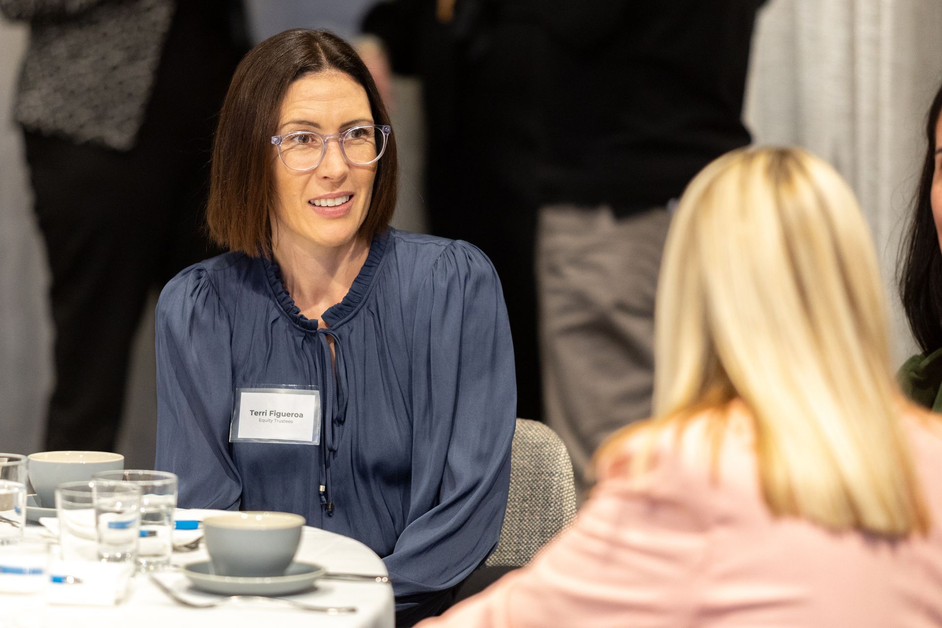 A woman wearing glasses is sitting at a table talking to another woman.