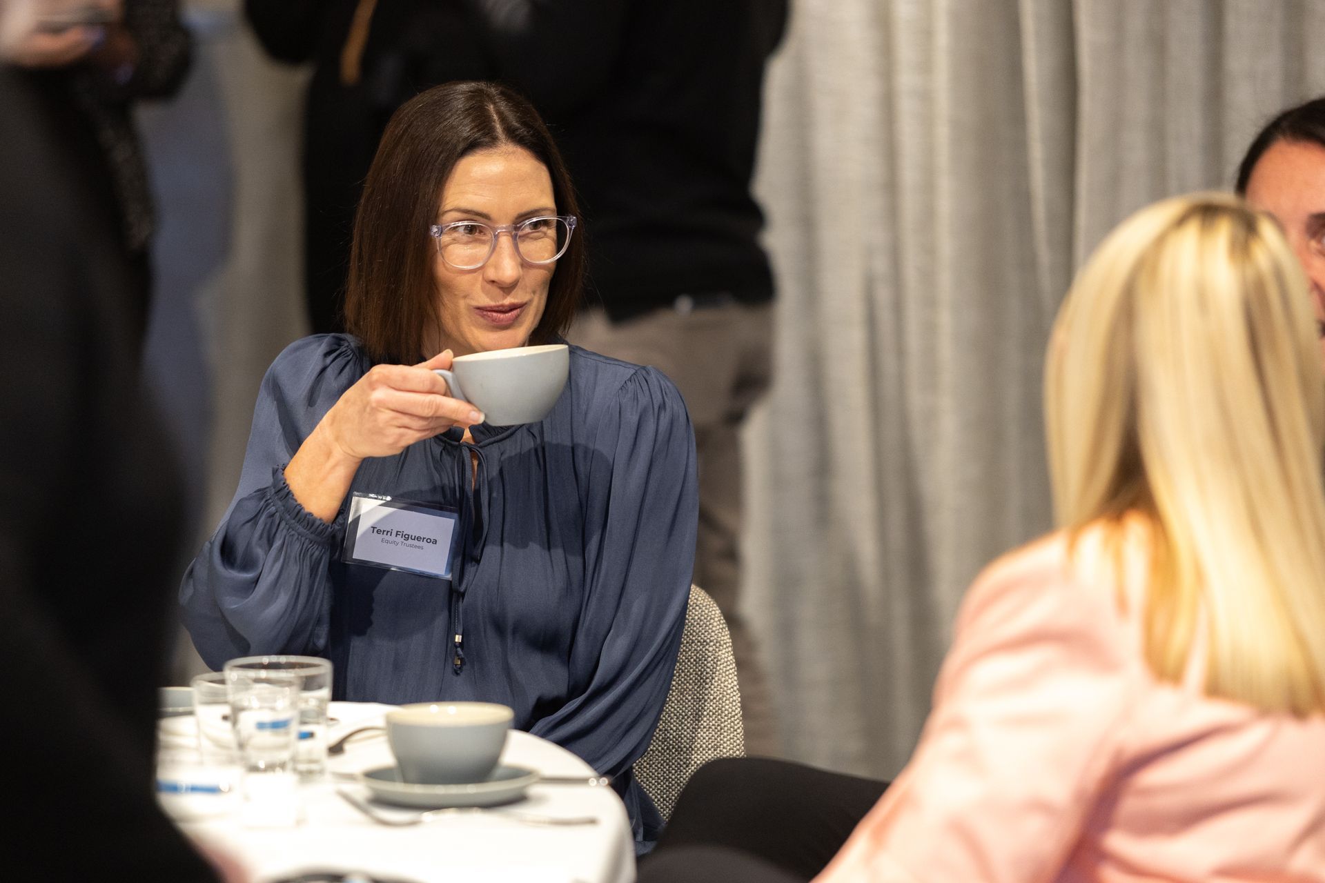 A woman is sitting at a table drinking a cup of coffee.