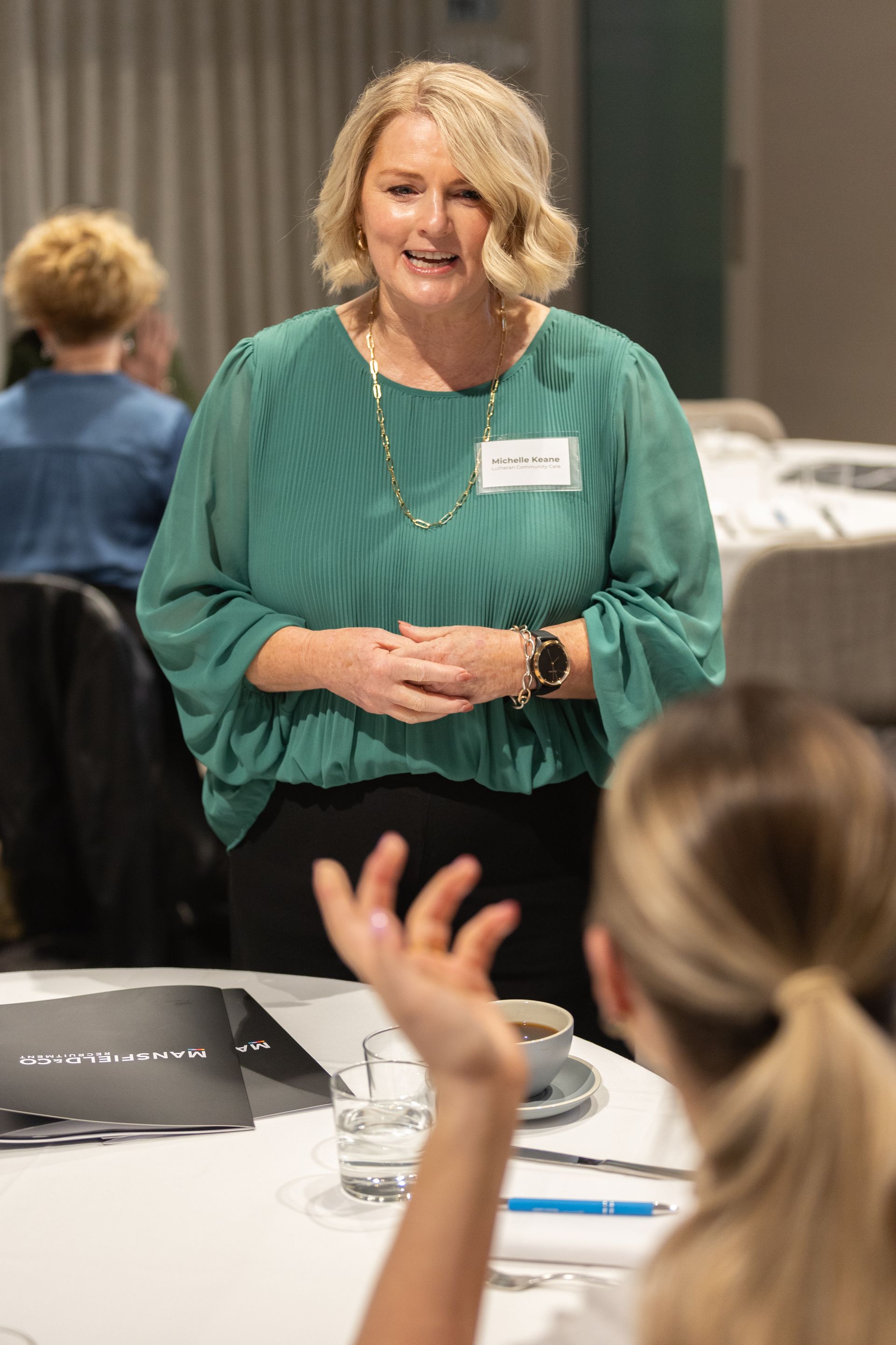 A woman in a green shirt is standing at a table talking to a group of people.