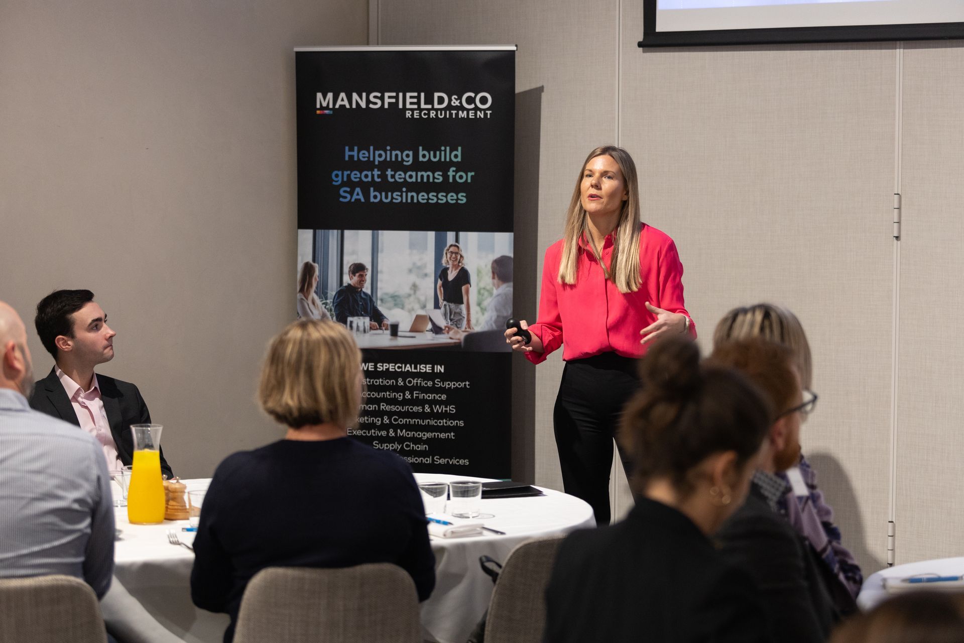 A woman is giving a presentation to a group of people sitting at tables.