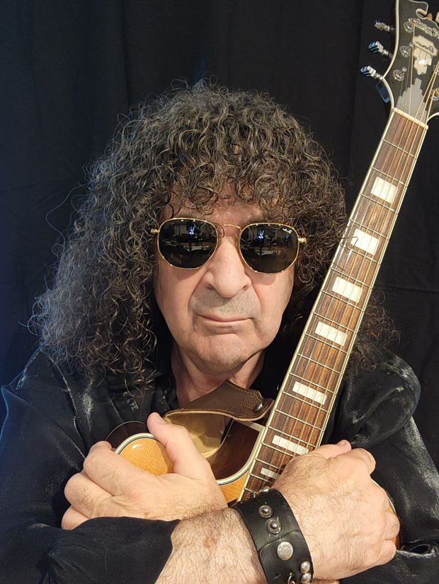 A man with curly hair and sunglasses is holding a guitar