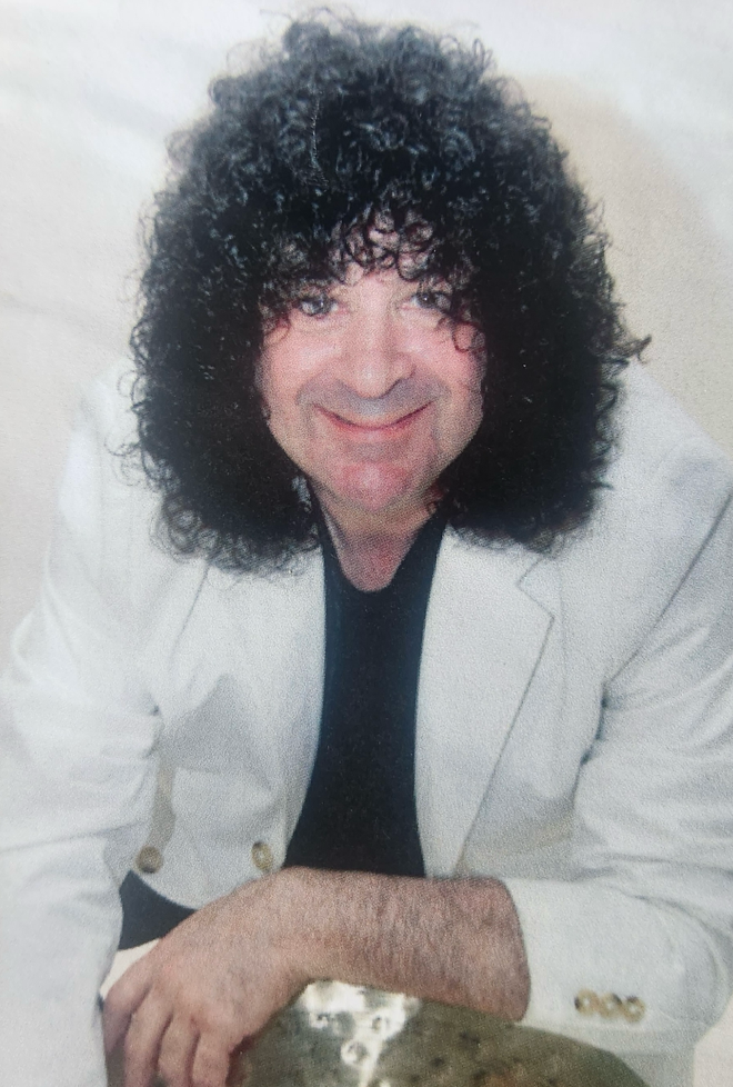 A man with curly hair and a white jacket is sitting next to a cymbal.