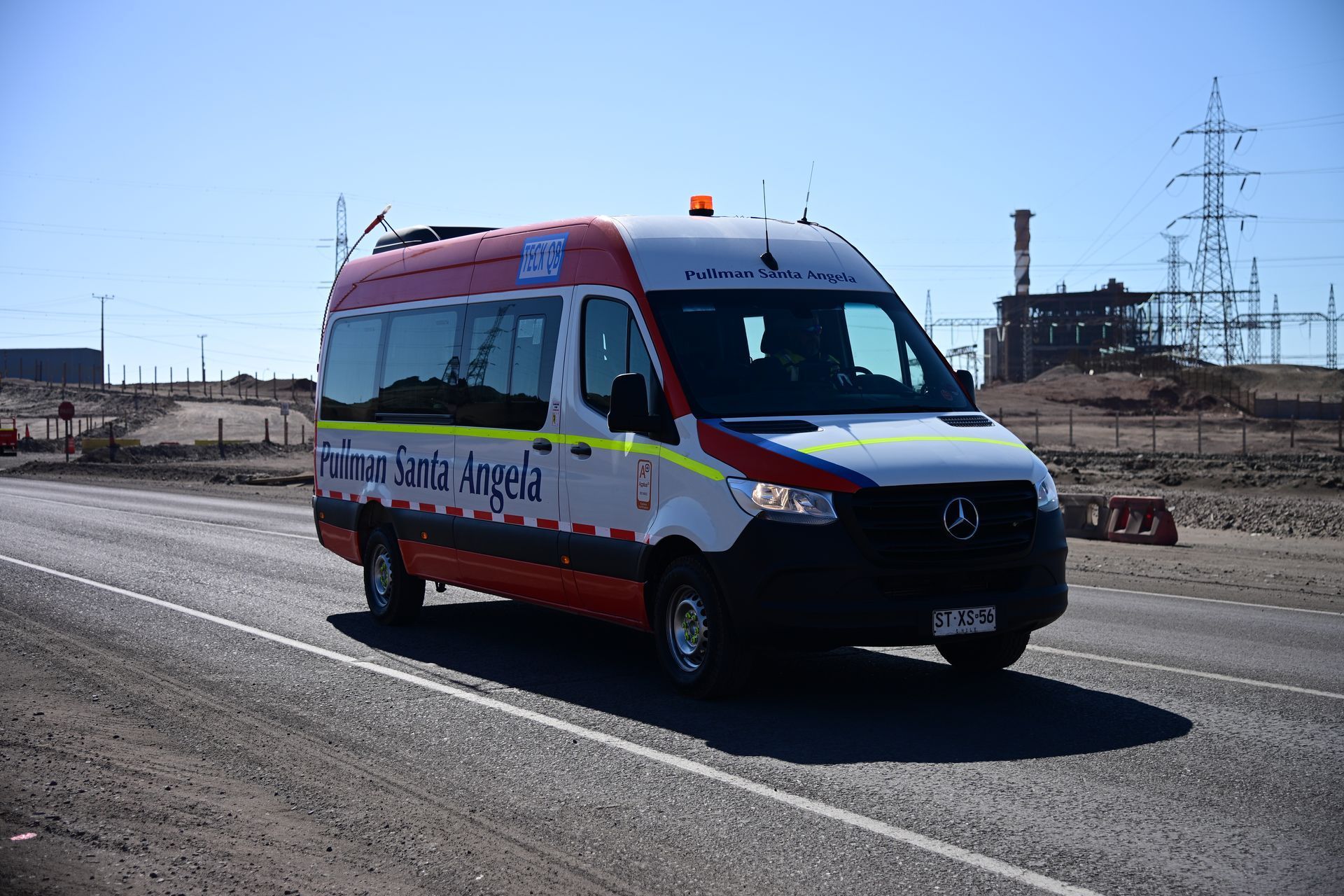 Ambulancia blanca, roja y negra en una carretera; planta eléctrica al fondo.