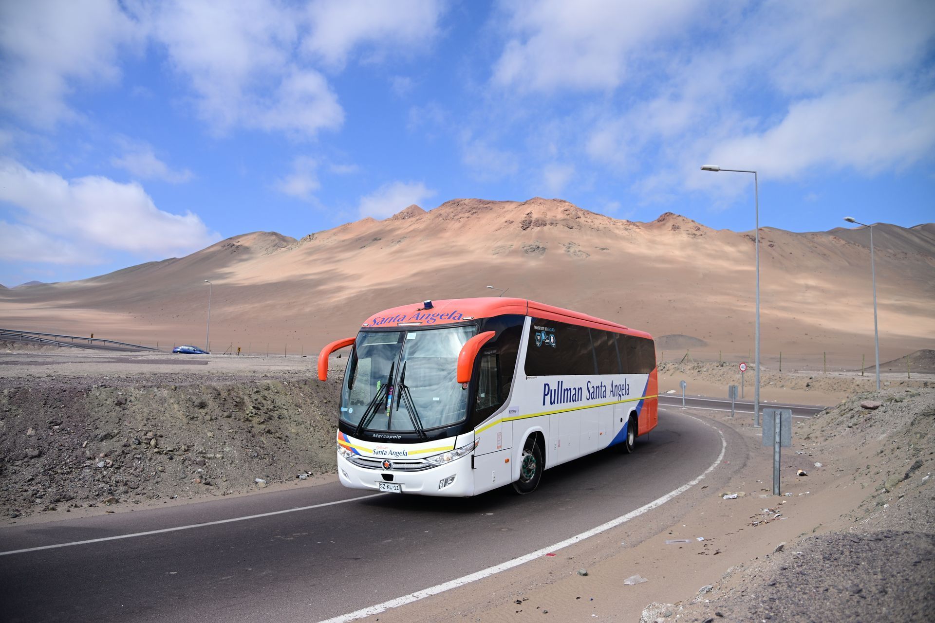 Autobús blanco y rojo circulando por una carretera desértica con montañas bajo un cielo azul.