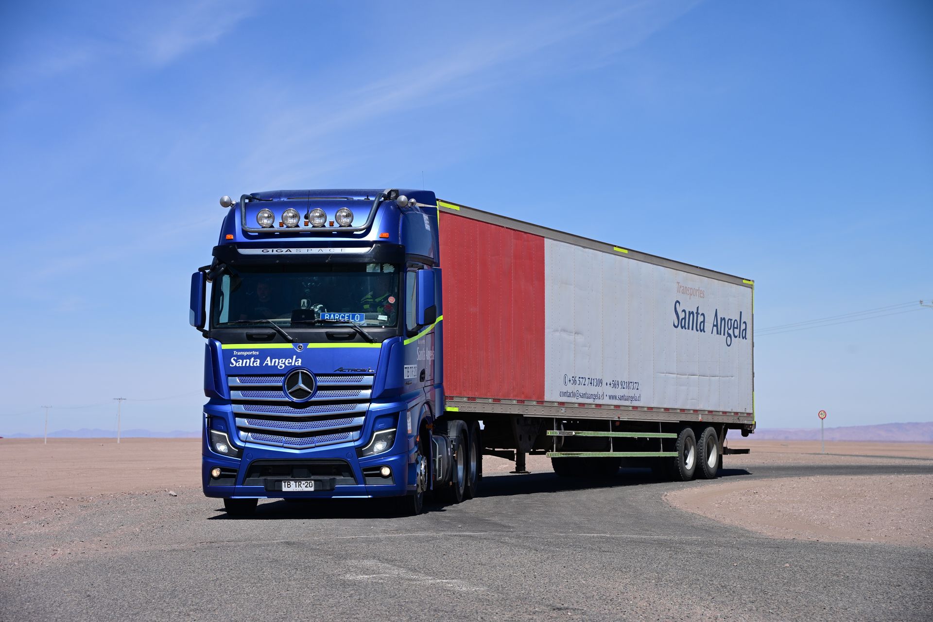 Autobús de pasajeros naranja y azul estacionado en un lote de grava bajo un cielo azul claro.