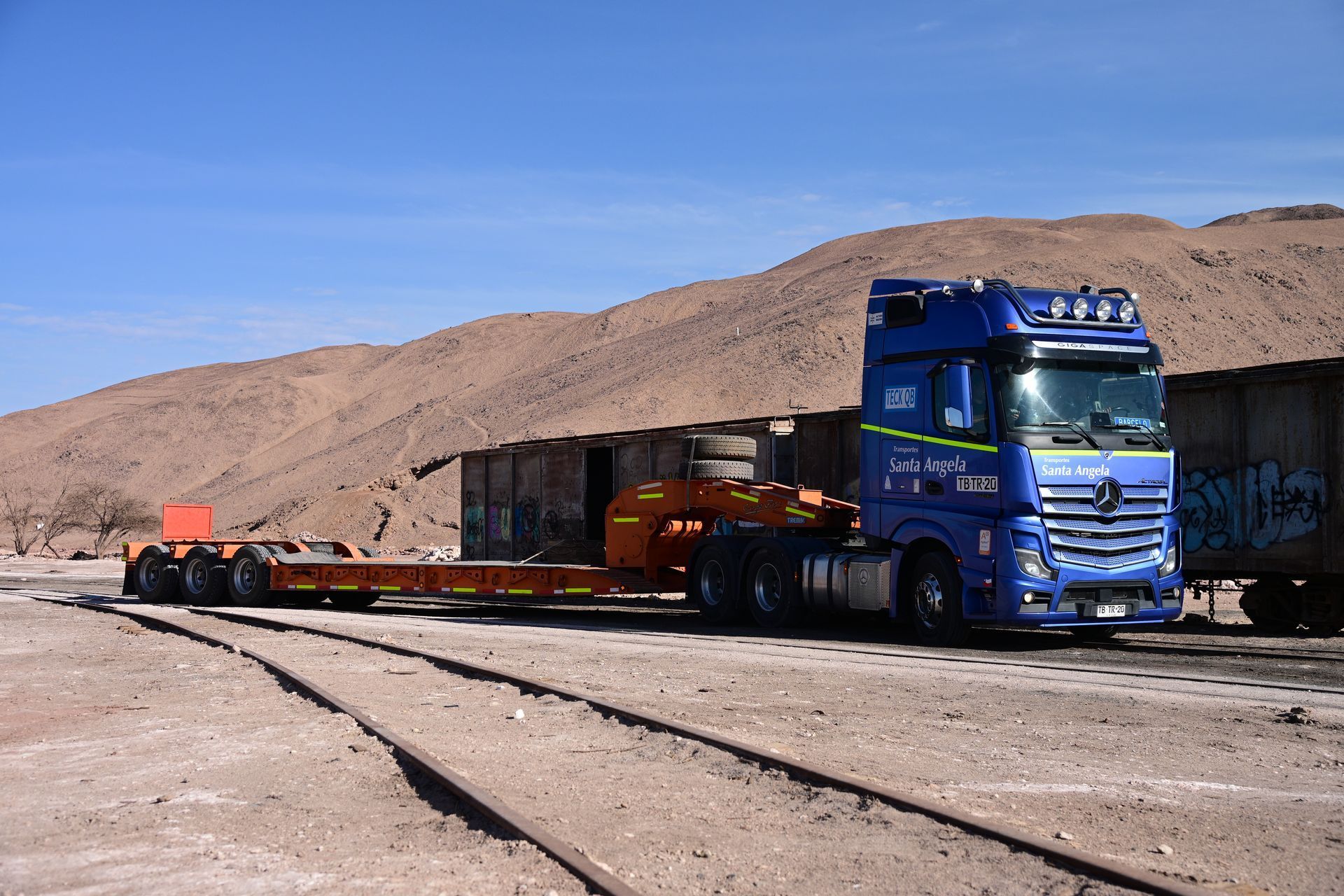 Camión semirremolque azul con remolque de plataforma sobre vías de tren, colinas marrones al fondo.