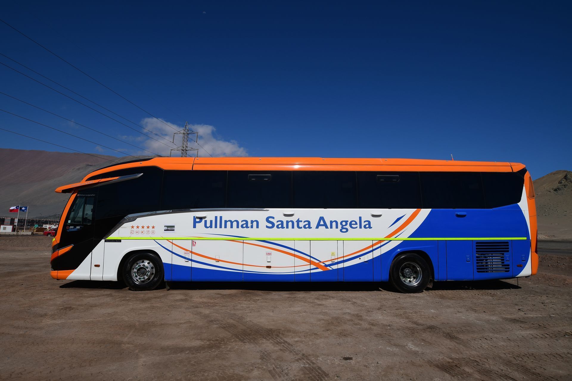 Autobús Pullman Santa Angela de color naranja, azul y blanco en un estacionamiento polvoriento al aire libre bajo un cielo azul.