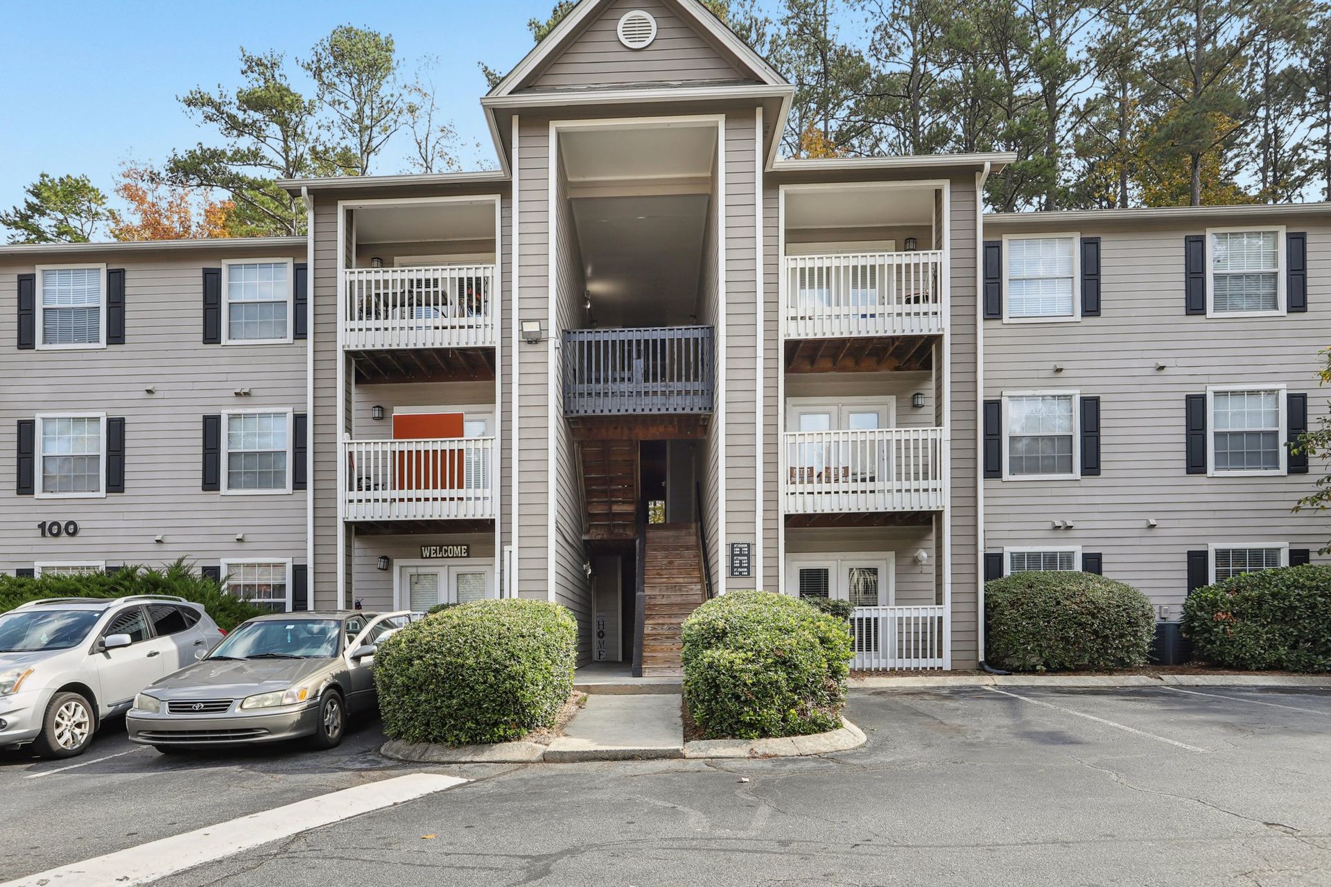 A three-story apartment building with beige siding, white balconies, and parking in front.