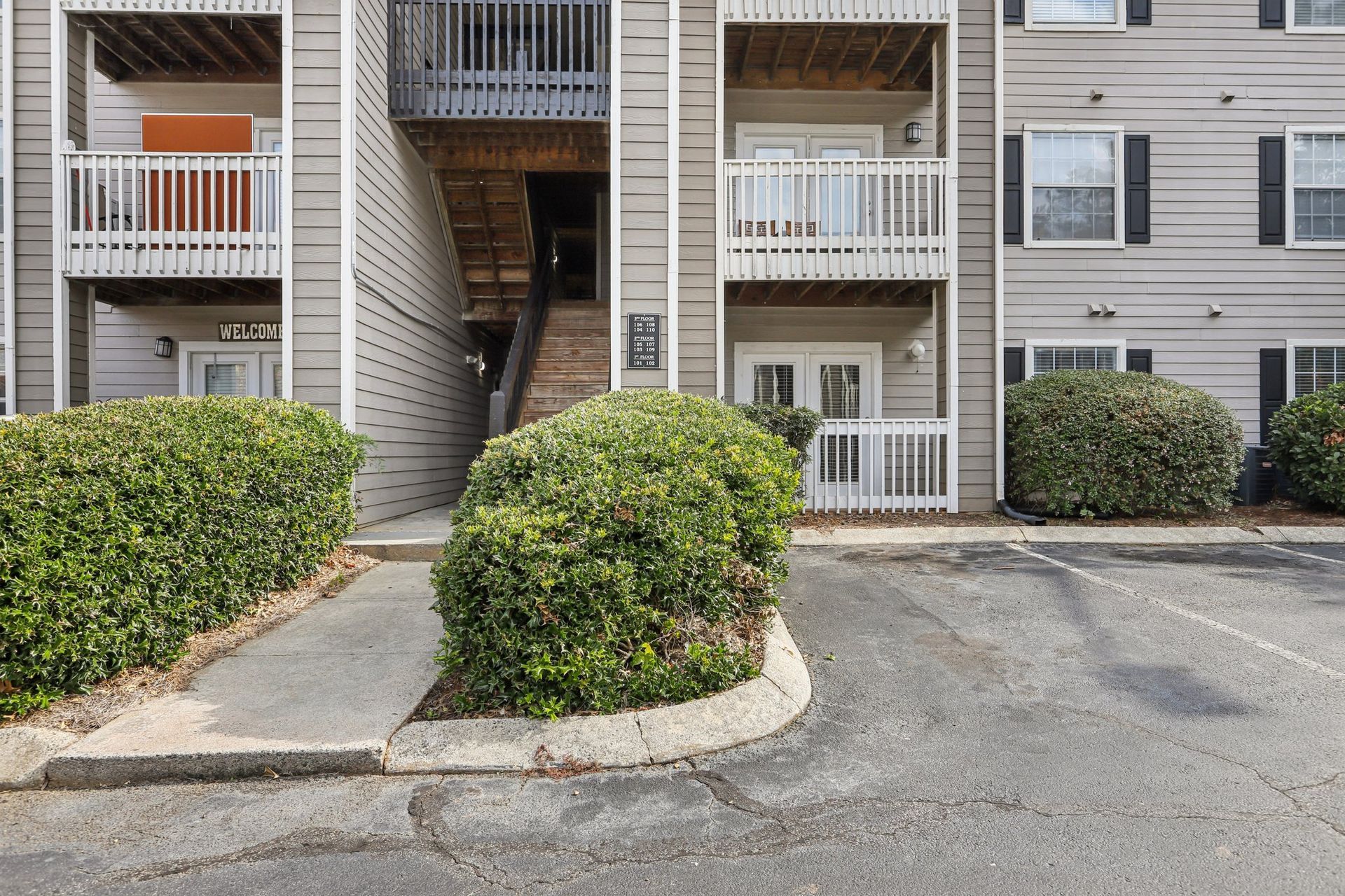 A multi-story apartment building with beige siding, white railings, and a stairwell entrance, fronted by bushes and parking.