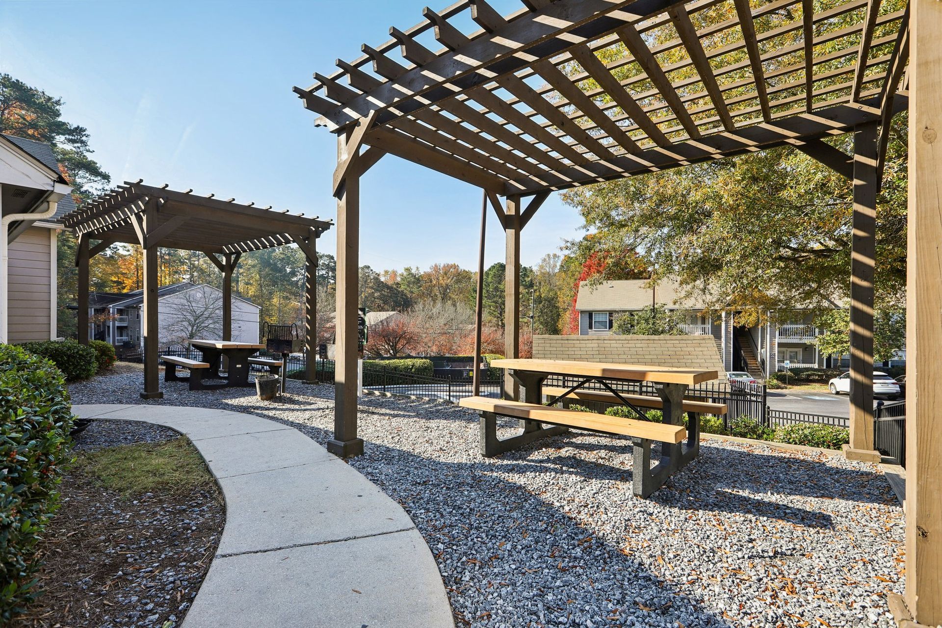 Two wooden pergolas with picnic tables stand over a gravel patio with a curved concrete walkway on a sunny day.