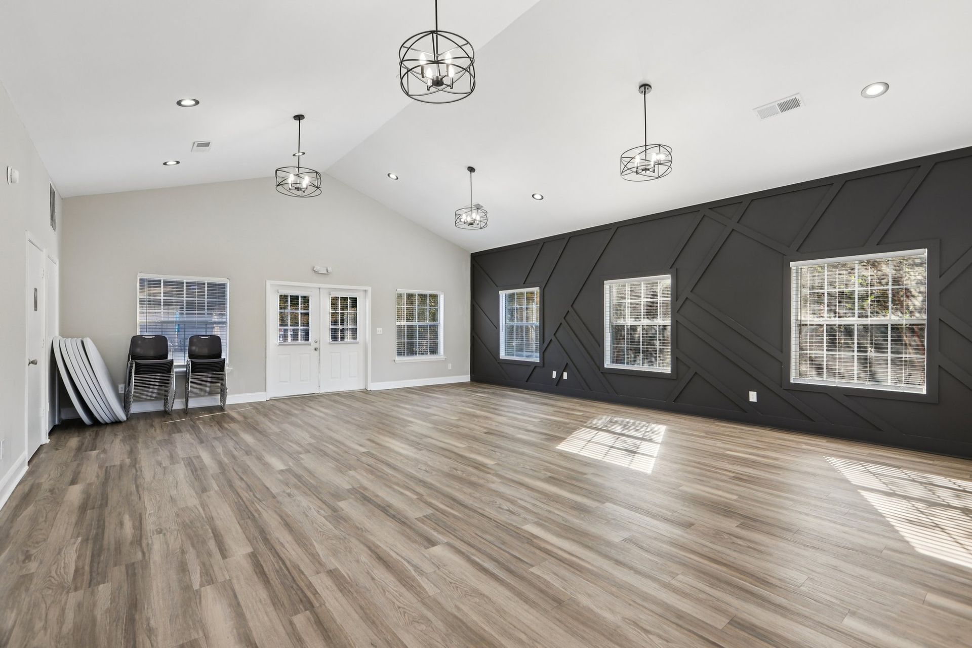 Empty community room with wood floors, vaulted ceiling, black accent wall with geometric trim, and hanging lights.