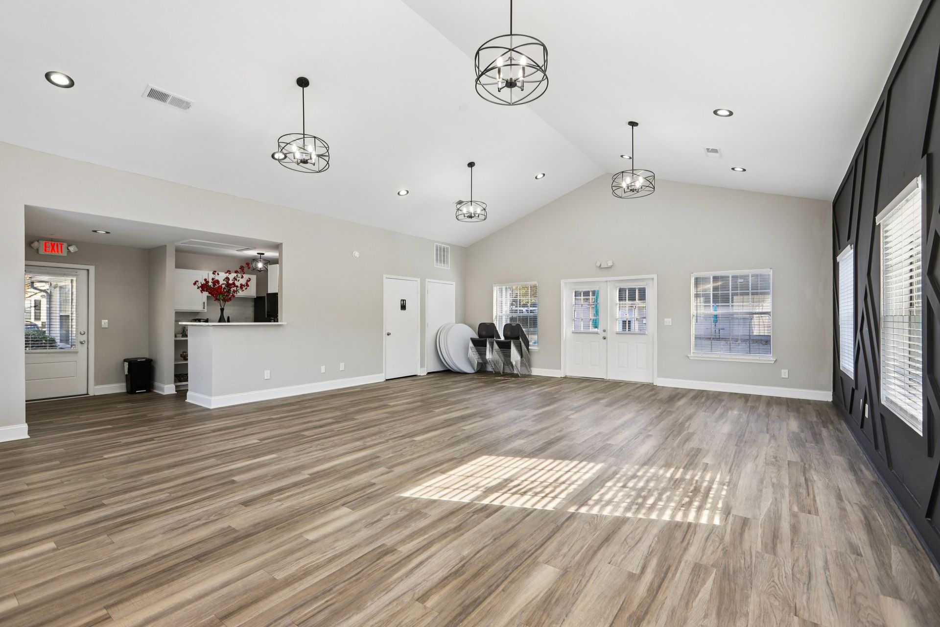 A bright, open community room with wood-look flooring, light grey walls, white doors, and modern pendant lighting.