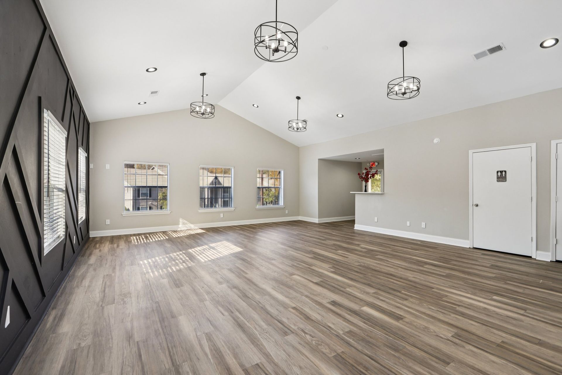 A bright, empty event room with light wood floors, a dark patterned accent wall, three windows, and hanging light fixtures.