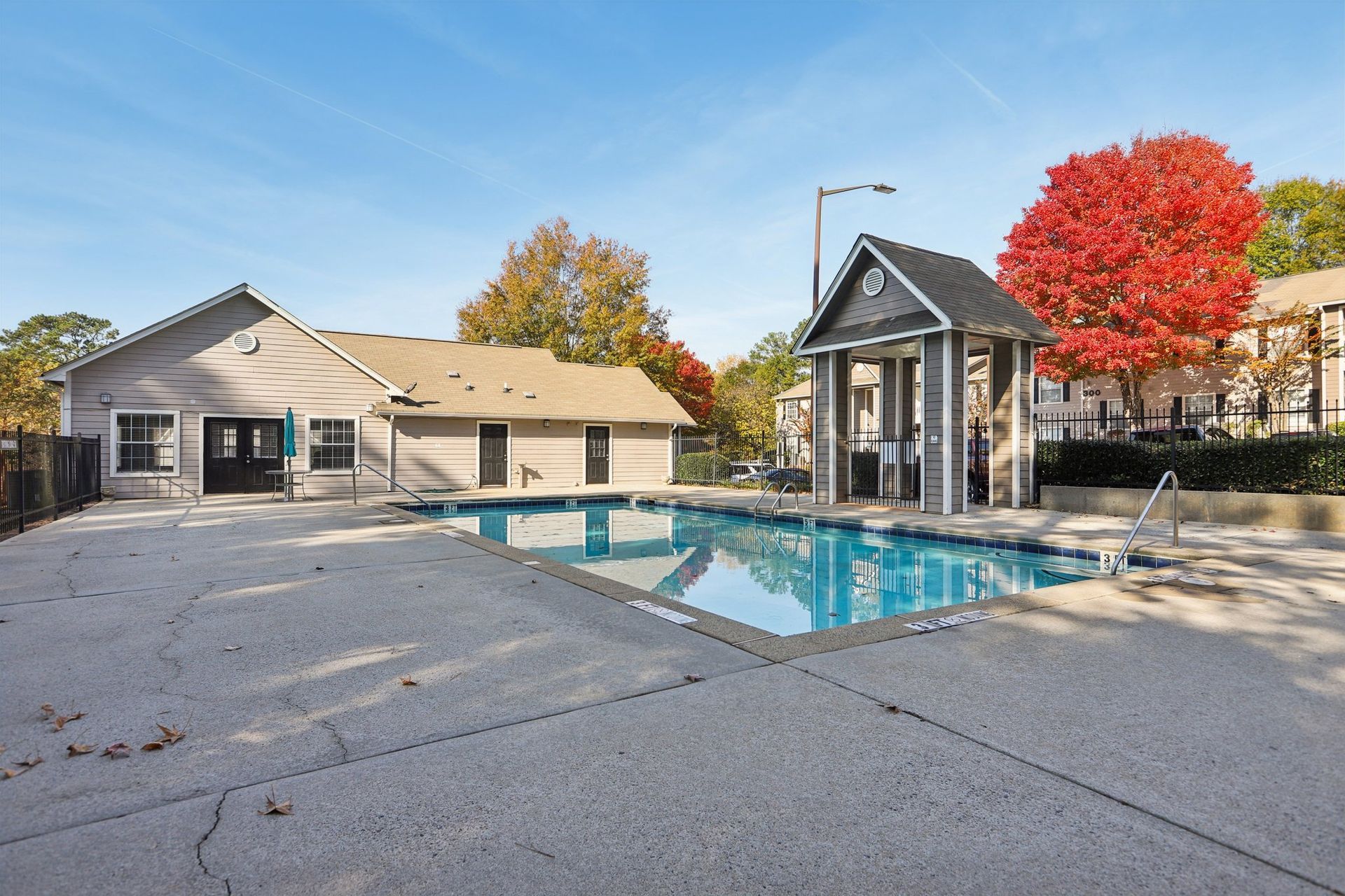 A rectangular outdoor swimming pool next to a one-story building and a covered gazebo on a sunny day with autumn foliage.