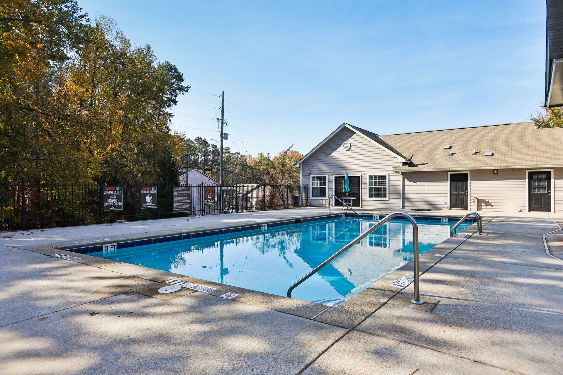 A swimming pool with blue water sits in front of a single-story building under a clear blue sky, surrounded by trees.