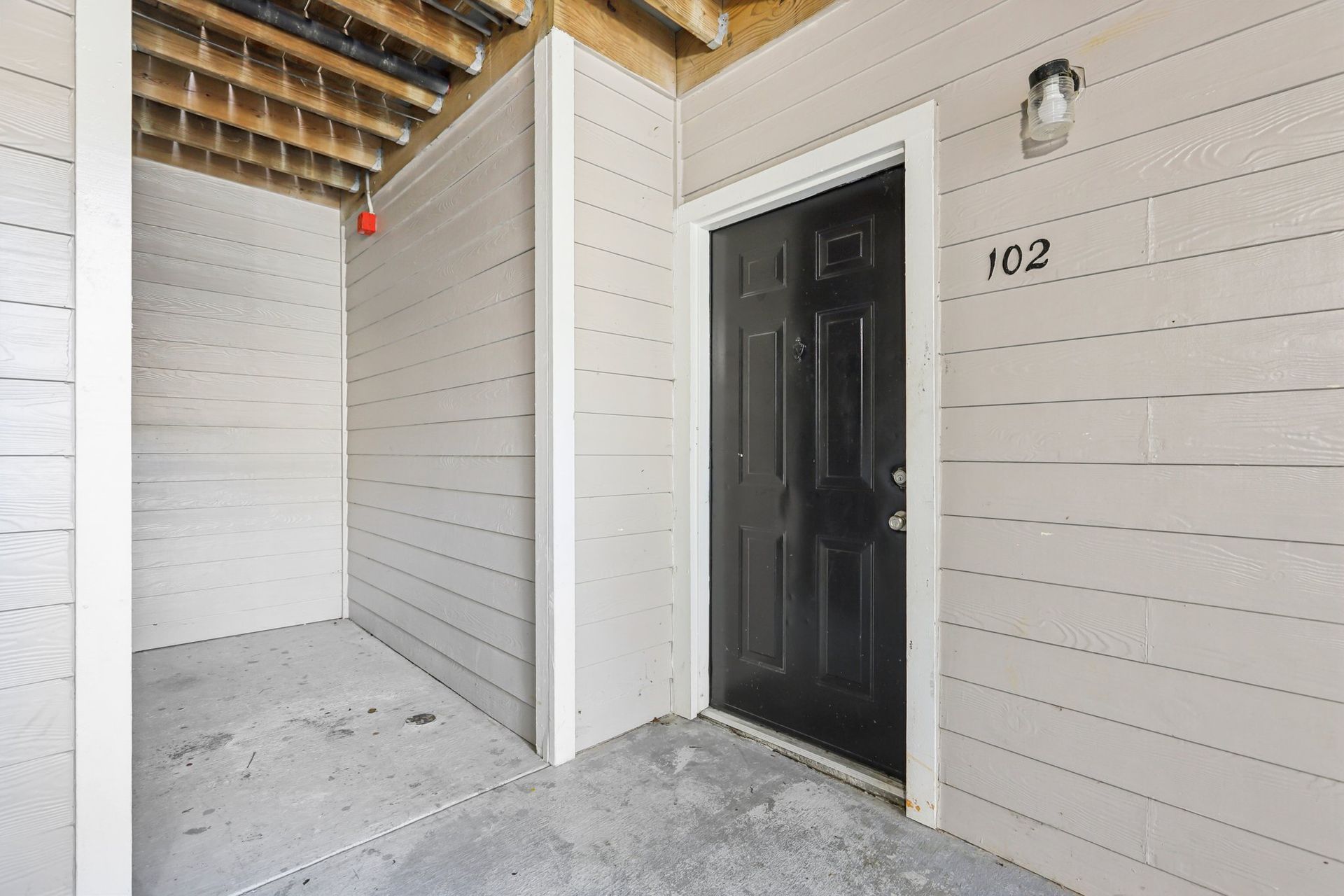 A dark door labeled 102 under a covered walkway with light-colored horizontal siding and a concrete floor.