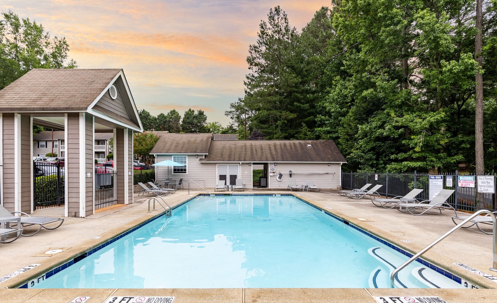 A swimming pool with blue water sits in front of a single-story building under a clear blue sky, surrounded by trees.