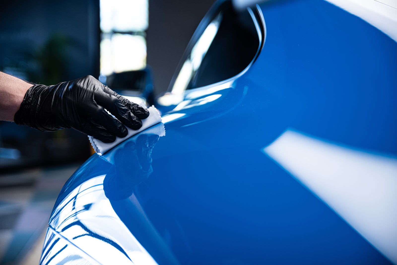 A hand in a black glove applies a protective coating to a shiny, bright blue car panel.