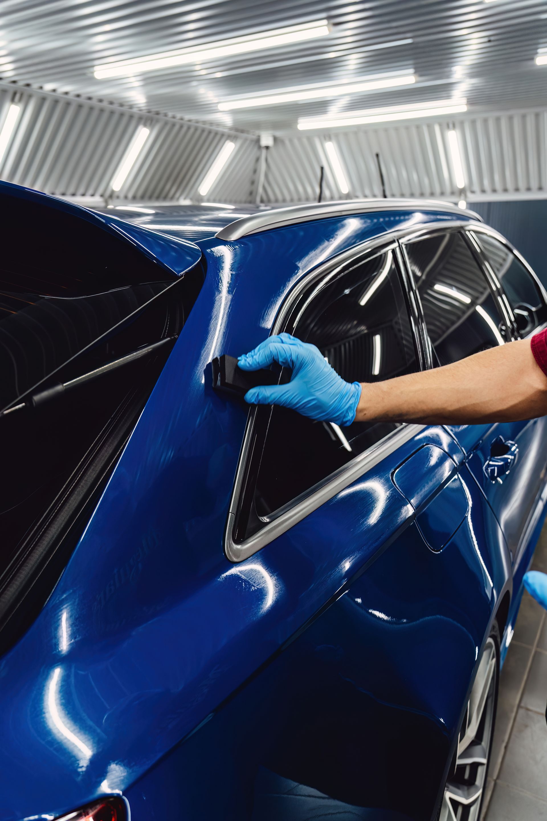A hand in a blue glove uses a small applicator to apply ceramic coating to the blue paint of a car in a detailing shop.