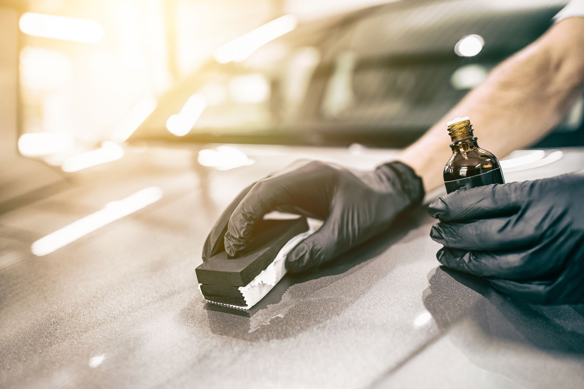 A person wearing black gloves applies a protective ceramic coating to a car hood using a small applicator block.