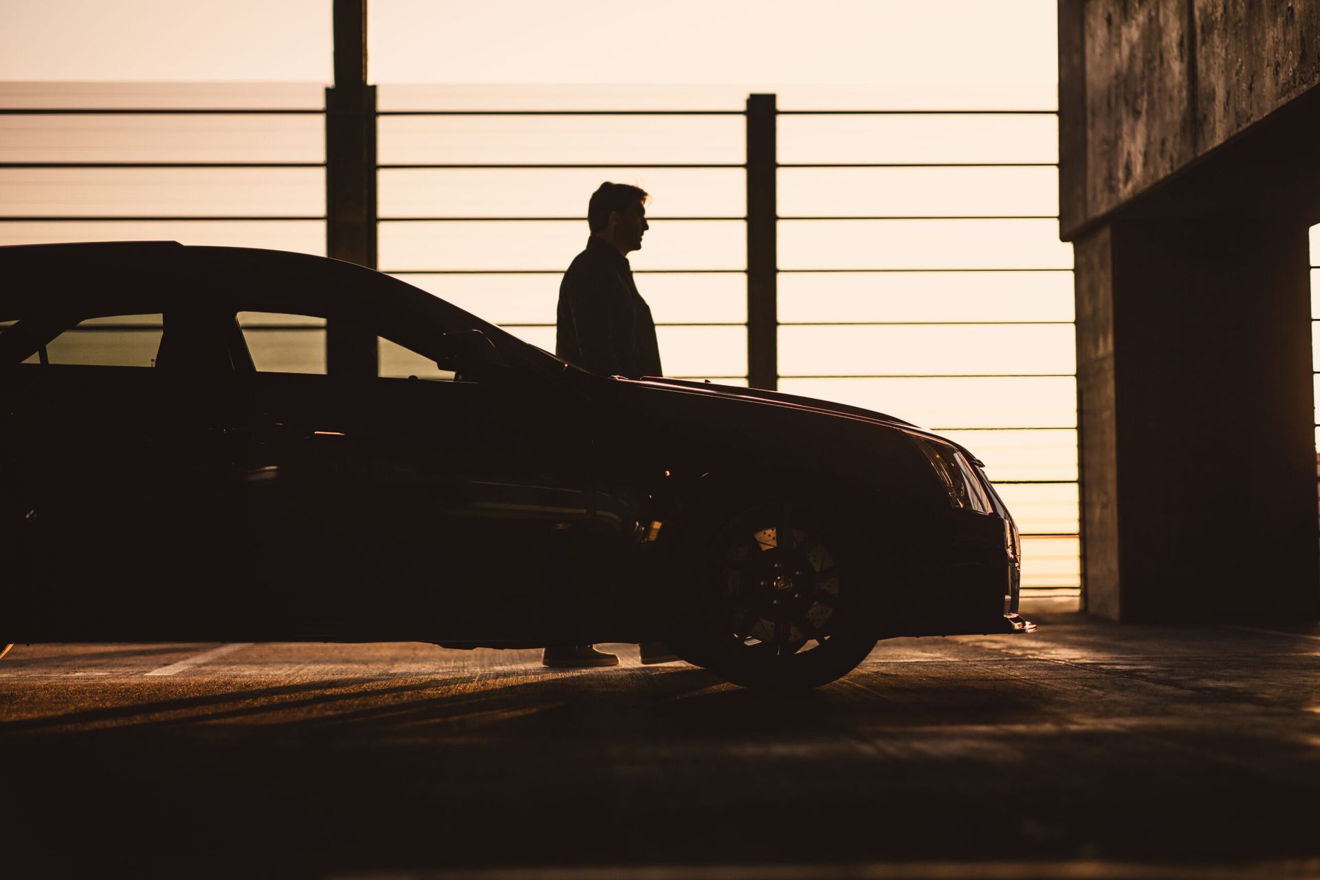 A silhouette of a person standing next to a car in a parking garage at sunset.