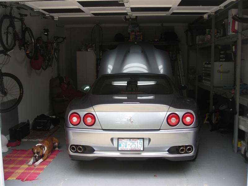 A silver Ferrari parked in a garage with its trunk open, next to a dog resting on a mat and bicycles hanging on the wall.