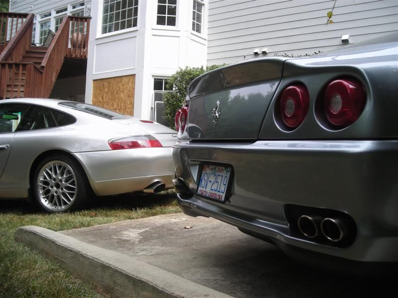 A silver Porsche parked in a grassy area next to a grey Ferrari with dual exhaust pipes parked on a concrete driveway.