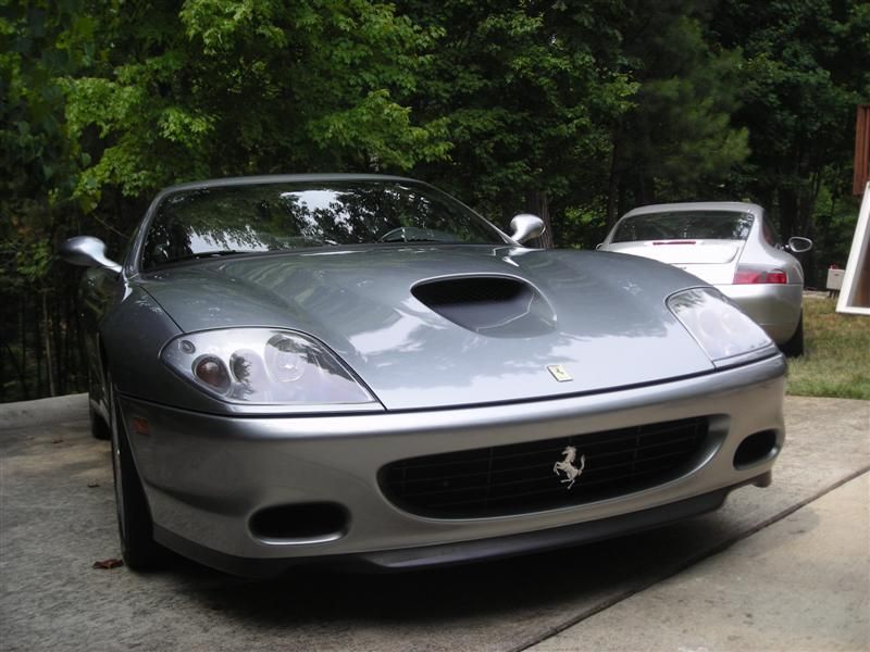 A silver Ferrari 575M Maranello parked on a driveway with a car partially visible in the background.