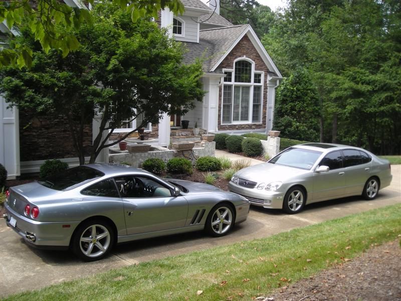 A silver sports car parked next to a silver sedan in the driveway of a brick house surrounded by trees.