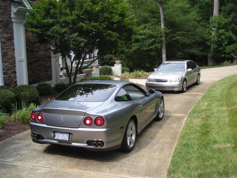 A silver Ferrari sports car parked in a driveway in front of a silver sedan near a house with lush green trees.