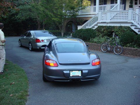 A gray Porsche and a silver sedan parked in a driveway in front of a house, with a bicycle leaning against a brick wall.