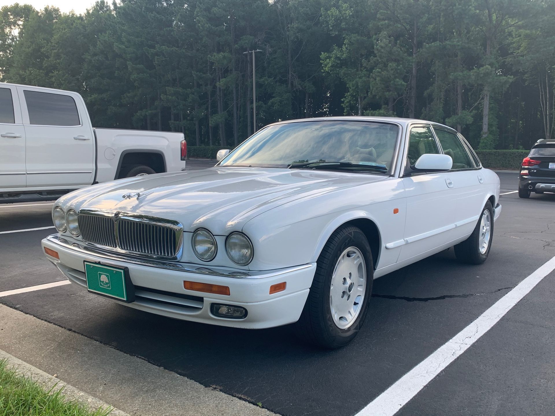 A white vintage Jaguar XJ sedan parked in a lot next to a white pickup truck on a sunny day.