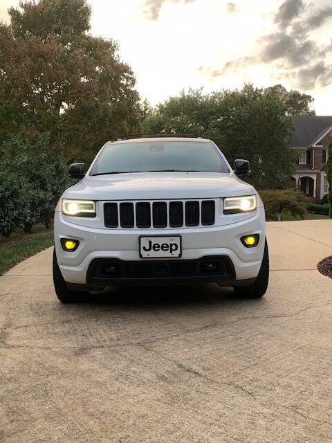 A white Jeep Grand Cherokee parked on a concrete driveway with yellow fog lights illuminated at dusk.