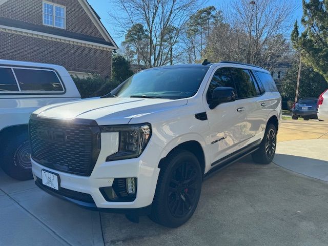 A white GMC Yukon SUV with a black grille and black wheels parked on a driveway in front of a brick house.