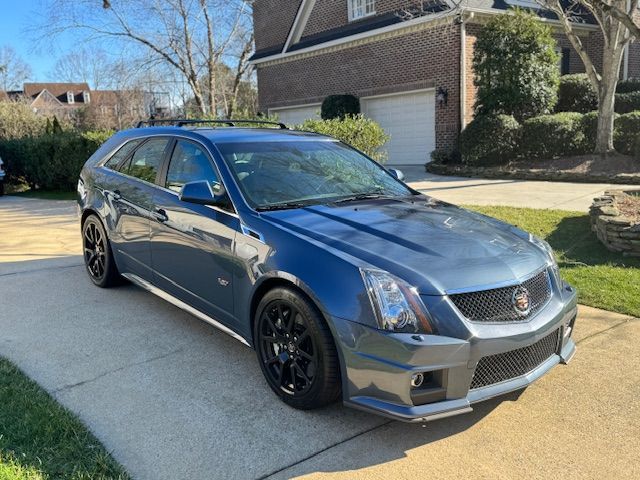 A blue Cadillac CTS-V wagon parked on a suburban concrete driveway in front of a brick house on a sunny day.