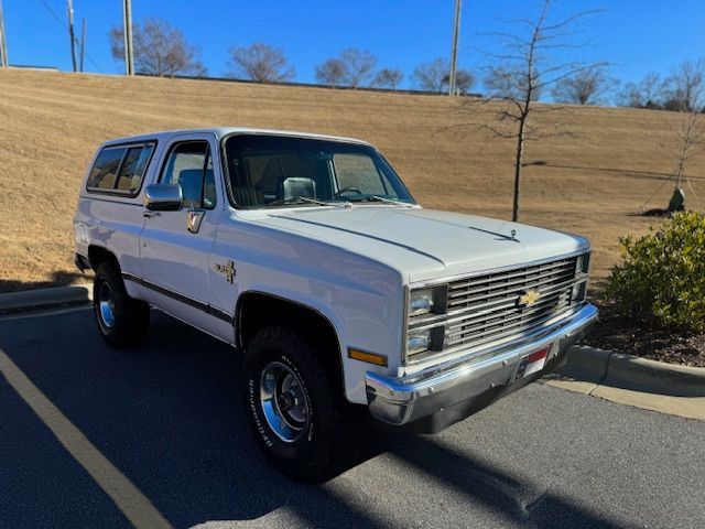 A white vintage Chevrolet K5 Blazer parked on asphalt in front of a grassy hillside under a clear blue sky.