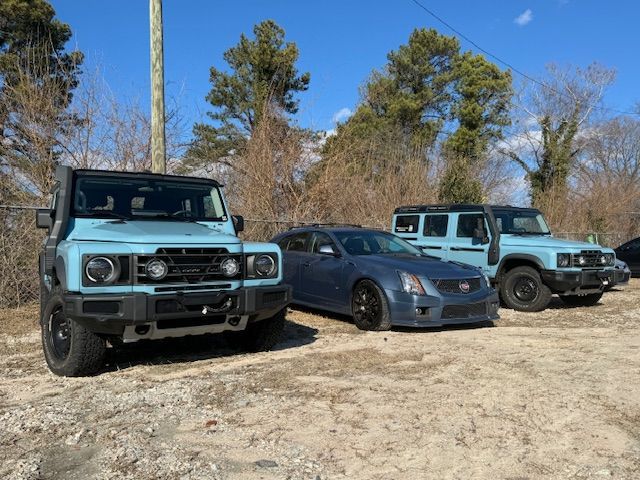 Two light blue INEOS Grenadier SUVs parked on either side of a dark blue Cadillac sedan on a gravel lot under a clear sky.